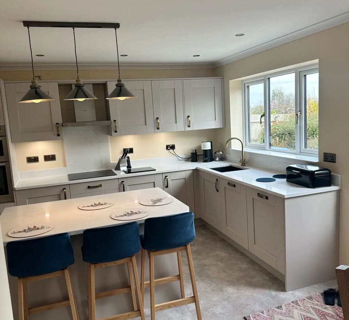 A modern kitchen with light gray cabinets, white countertops, and stainless steel appliances. There is a kitchen island with three blue bar stools, pendant lights, and a window overlooking a garden.